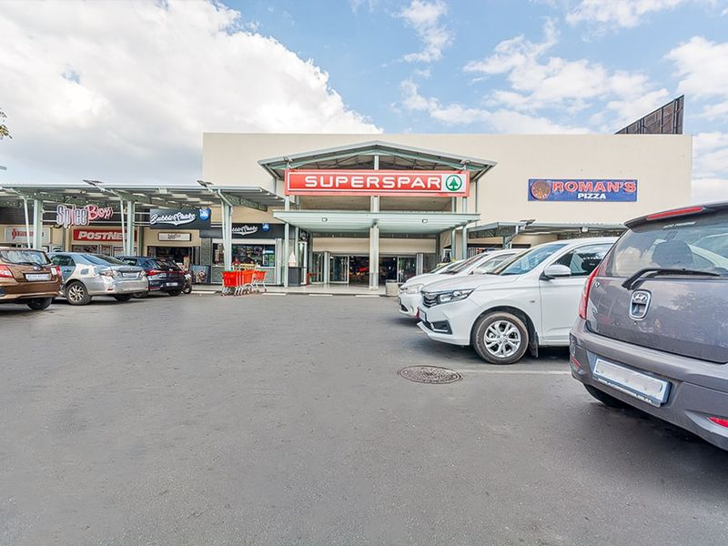 Front exterior view of a commercial shopping center featuring a SuperSpar anchor store, various retail storefronts, and an asphalt parking area.