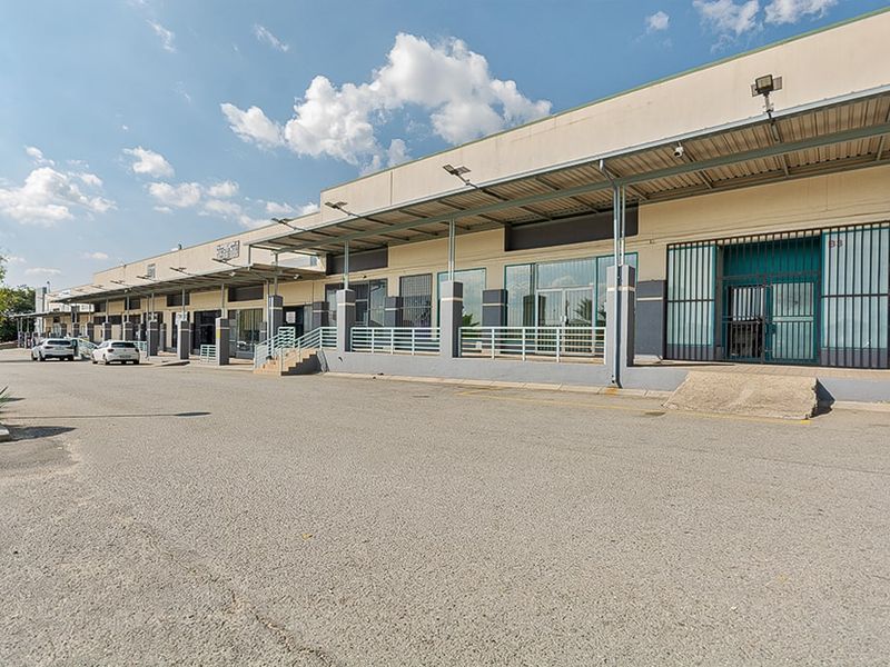 Commercial property exterior featuring a row of storefronts with metal canopy coverage, security gates, and an expansive asphalt parking area.