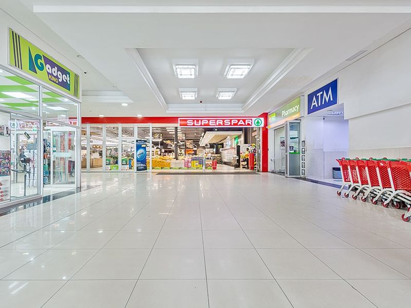 A bright, well-maintained commercial shopping mall corridor featuring tiled floors, recessed lighting, and multiple storefronts including a supermarket and pharmacy.