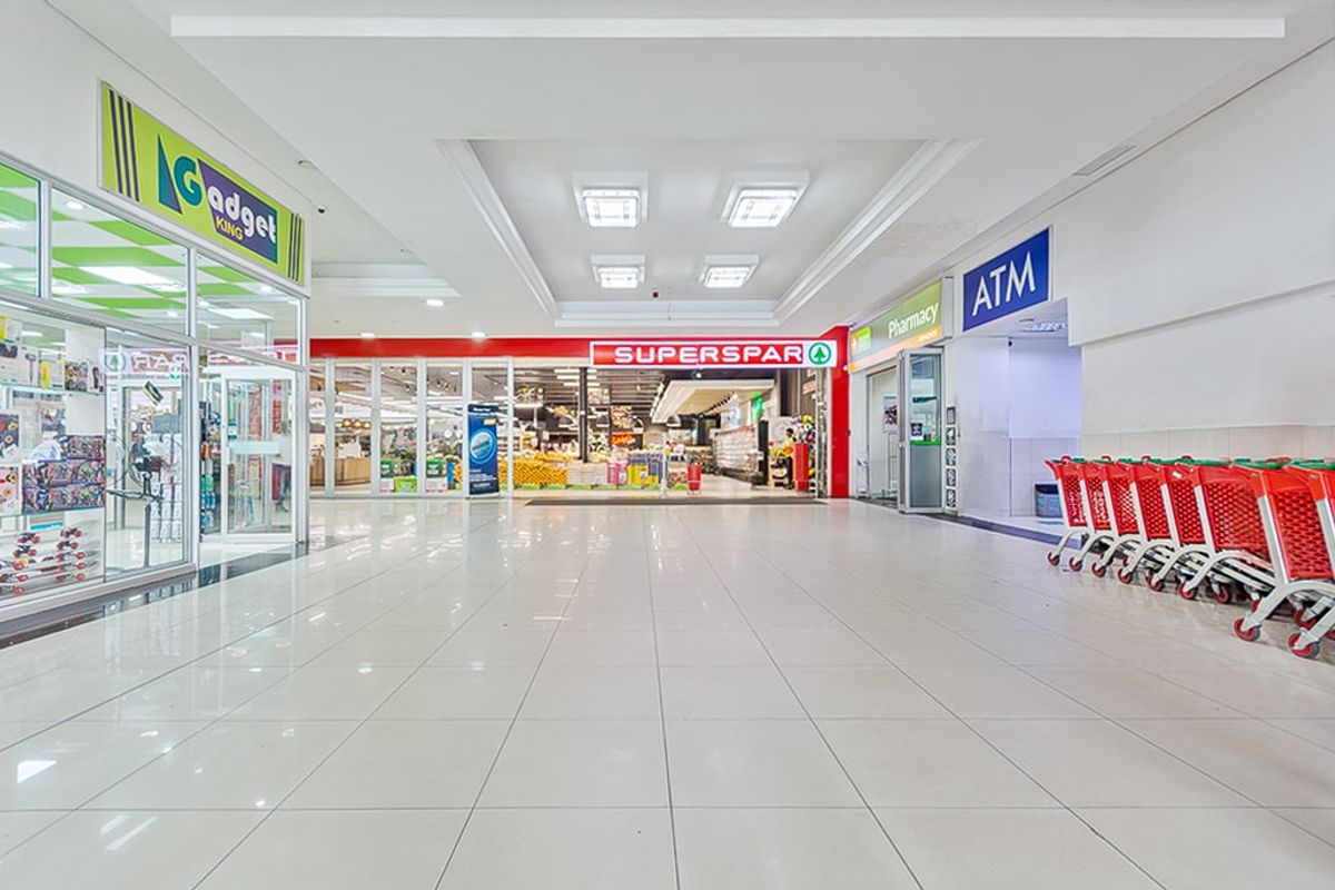 A bright, well-maintained commercial shopping mall corridor featuring tiled floors, recessed lighting, and multiple storefronts including a supermarket and pharmacy.
