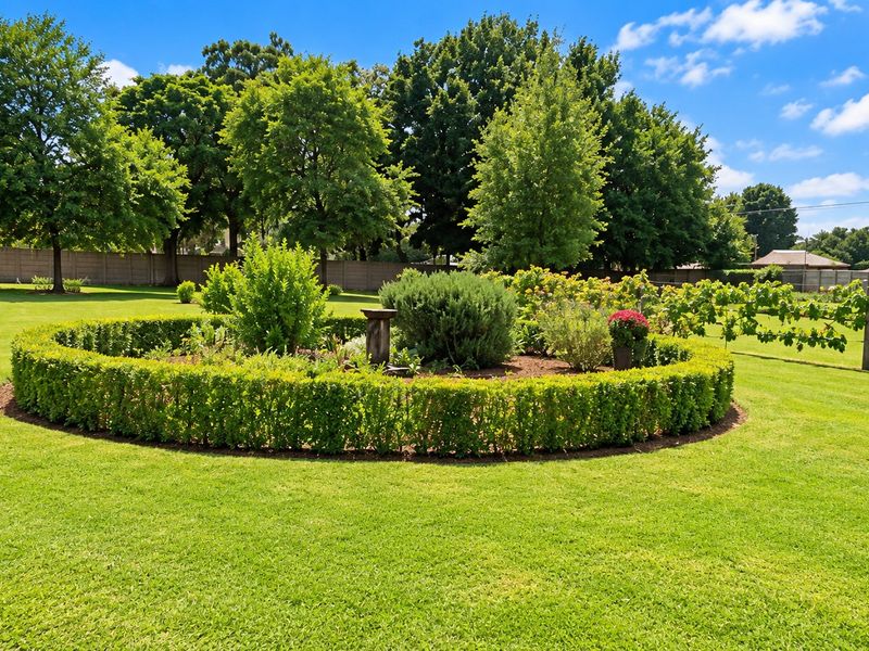 A well-maintained, expansive garden featuring a circular hedge bed, mature trees, and perimeter fencing.