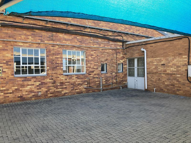 Rear exterior view of an industrial brick building featuring a paved courtyard area, security-fitted windows, and overhead shade sail protection.