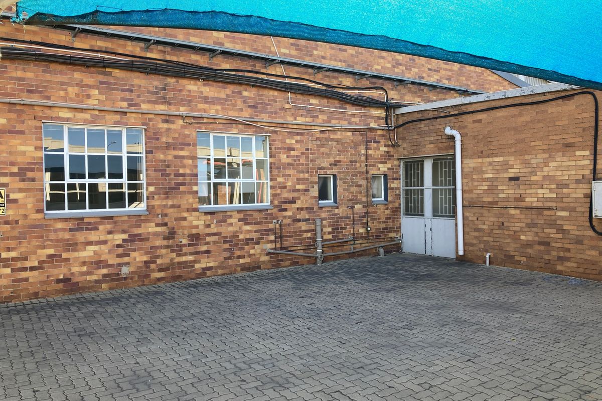 Rear exterior view of an industrial brick building featuring a paved courtyard area, security-fitted windows, and overhead shade sail protection.