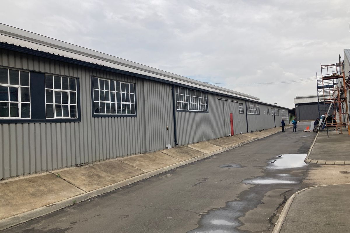 Exterior view of an industrial warehouse building featuring corrugated metal cladding, steel-framed windows, and an adjacent asphalt driveway with concrete curbing.