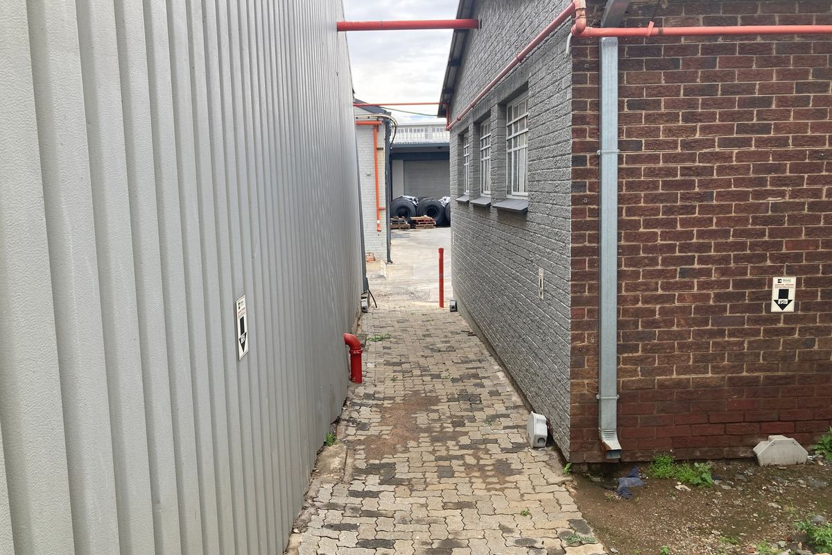 A narrow exterior walkway between industrial buildings featuring brick and corrugated metal walls with paved flooring and external plumbing infrastructure.