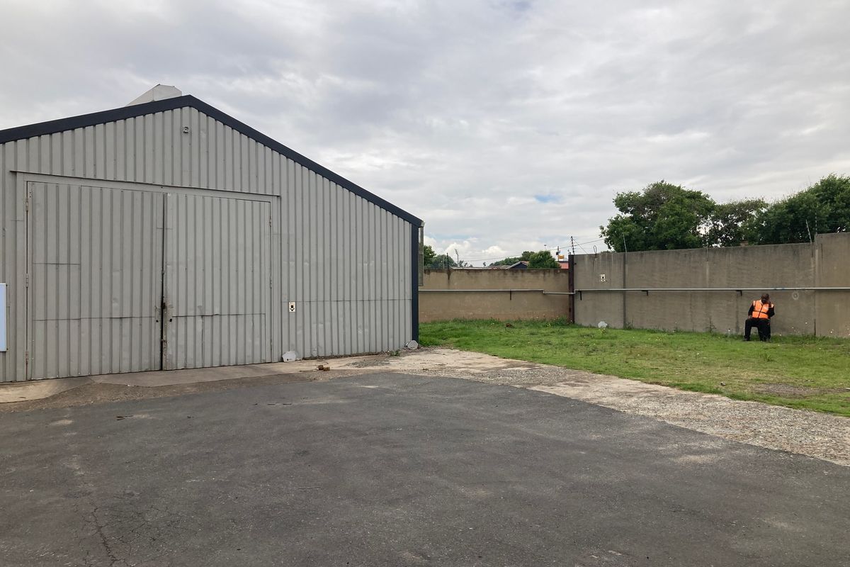 Rear industrial yard featuring a large corrugated metal warehouse building with sliding doors and a concrete perimeter wall.