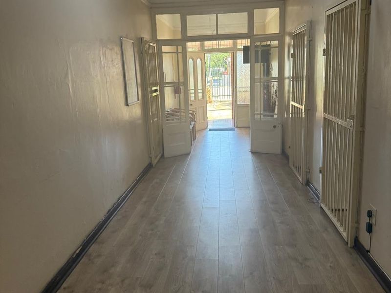 A long, vacant commercial hallway featuring laminate flooring, security gates, and glass-panelled doors leading to the building entrance.