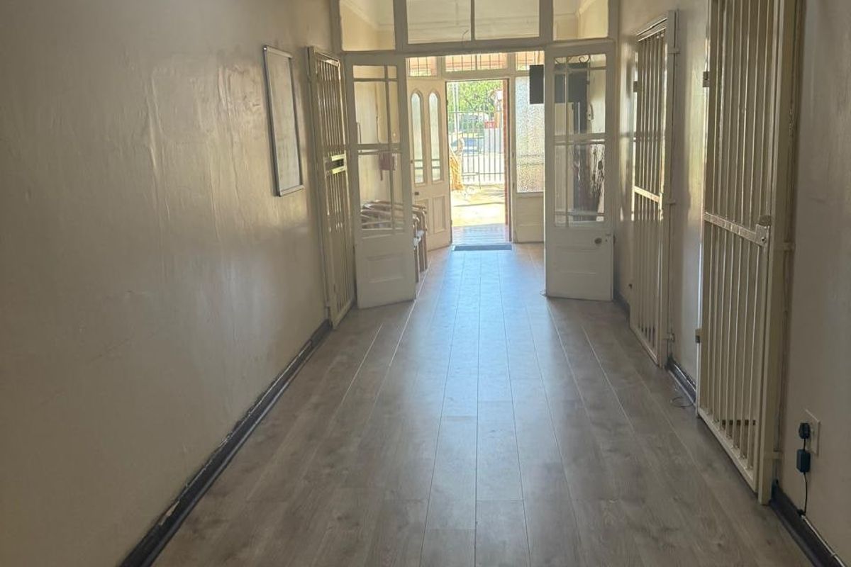 A long, vacant commercial hallway featuring laminate flooring, security gates, and glass-panelled doors leading to the building entrance.