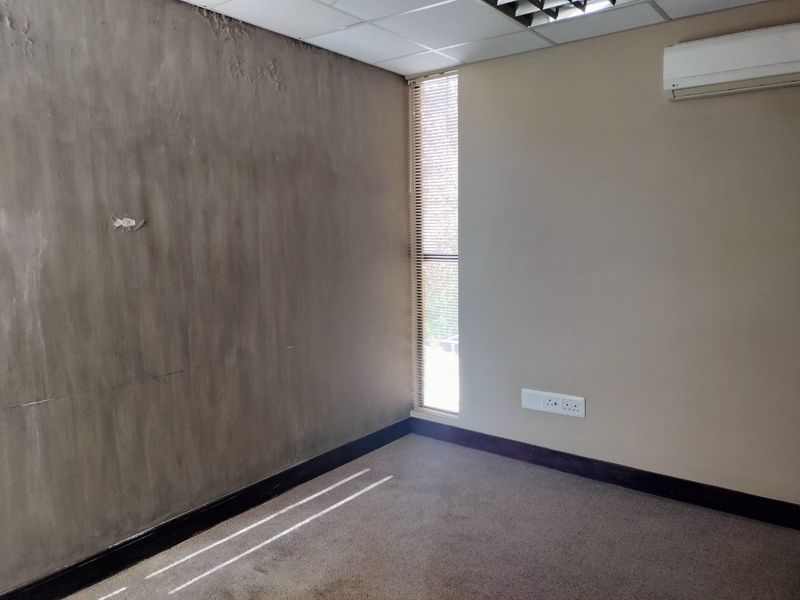 An empty office space featuring a suspended ceiling, wall-mounted air conditioning unit, and vertical window blinds, requiring cosmetic refurbishment to the walls and flooring.