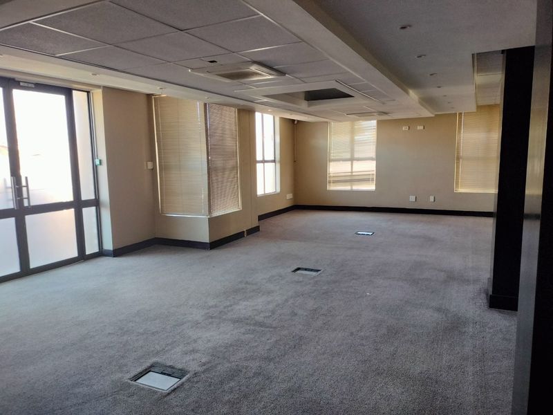 Spacious open-plan commercial office area featuring a suspended ceiling, recessed lighting, carpeted flooring with integrated floor power boxes, and large windows with venetian blinds.