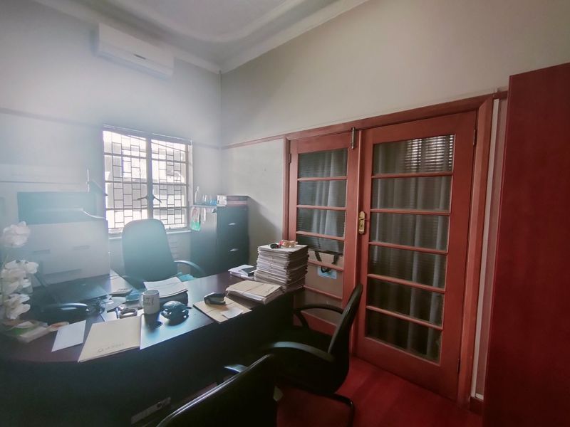 A functional office space featuring wooden double doors, a window with security bars, and a wall-mounted air conditioning unit.