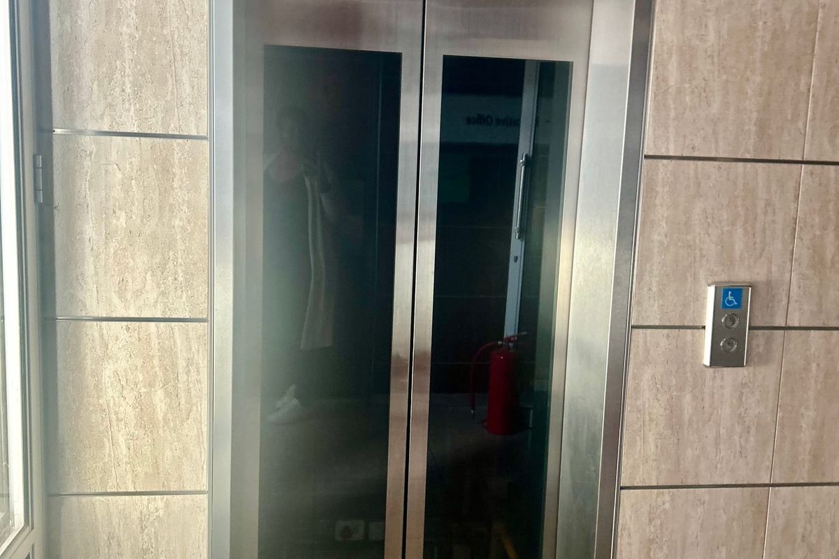 A clean commercial hallway featuring a modern stainless steel elevator with accessible call controls and neutral tiled wall finishes.