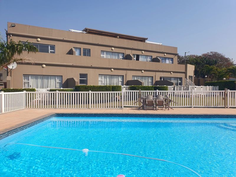 A well-maintained swimming pool area featuring a tiled deck, white picket fencing, and a view of the multi-story residential building facade.