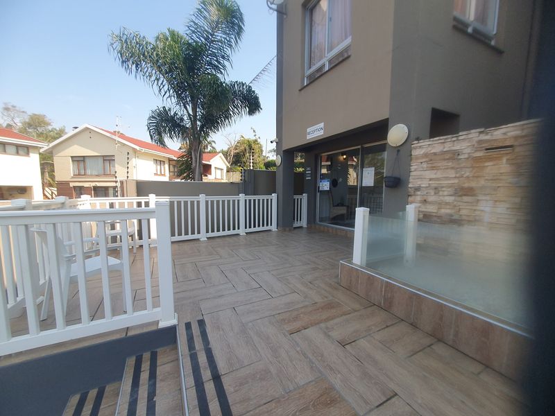 A well-maintained outdoor patio area featuring wood-look tiled flooring, a white balustrade, and a decorative stone feature wall near the building entrance.