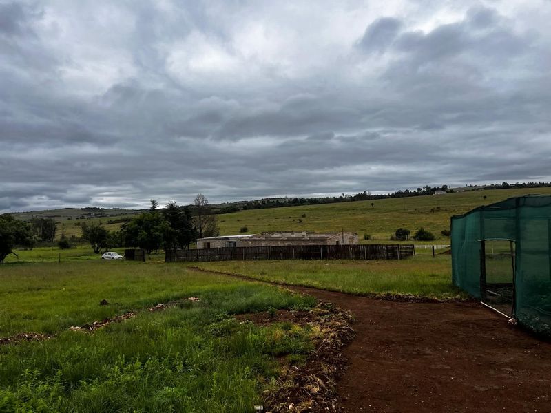 A wide view of a rural property featuring an unfinished building structure, perimeter fencing, and agricultural netting set against a grassy landscape.
