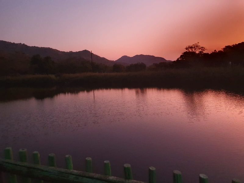 A scenic view of a tranquil dam at sunset, framed by a timber fence and rolling hills in the background.