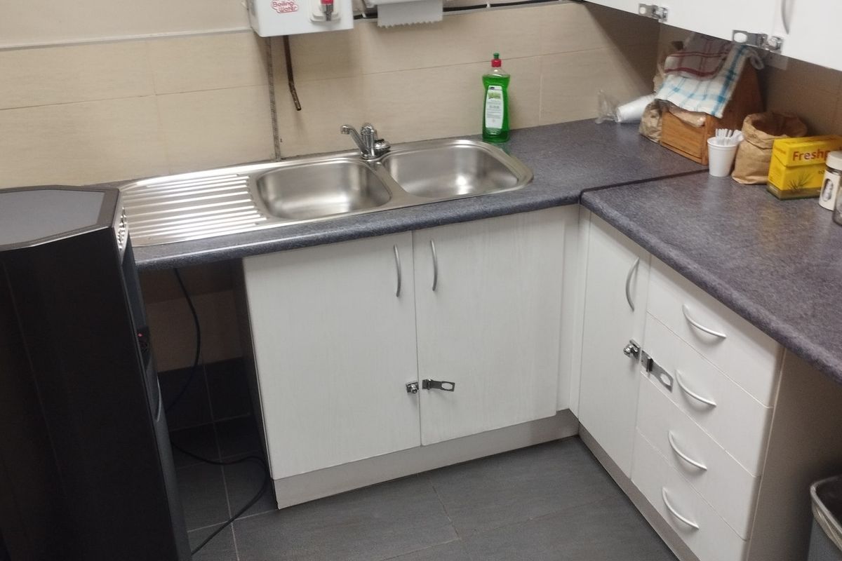 A functional kitchenette featuring white cabinetry, laminate countertops, and a double bowl stainless steel sink with wall-mounted water heater.