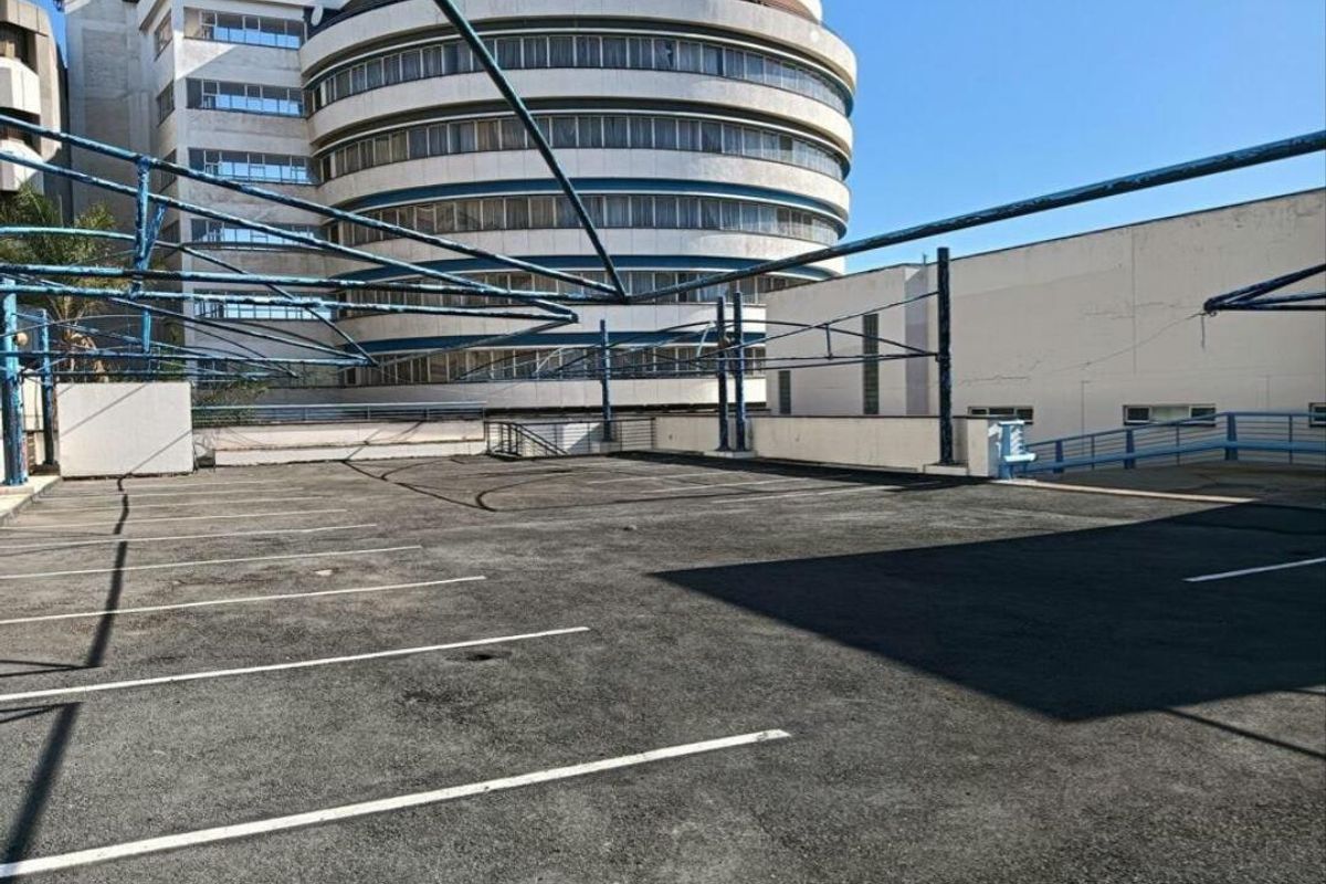 An open-air commercial parking area featuring an asphalt surface and a metal shade structure, set against a multi-story circular building.