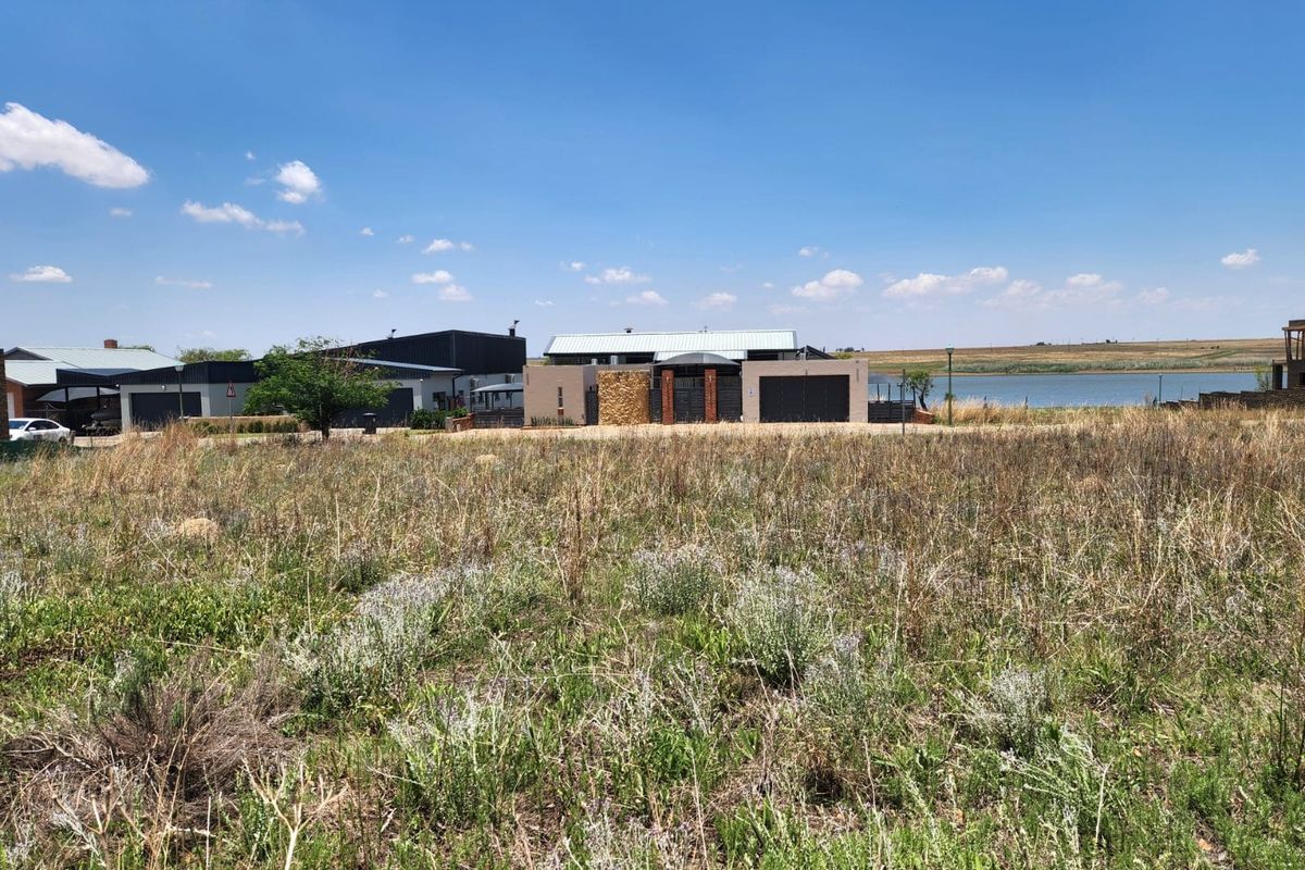 A view of a vacant residential plot with natural vegetation, situated in a developed estate near a water body.