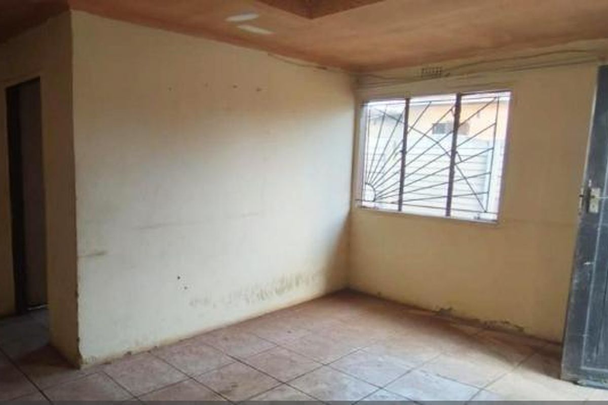 An empty living room featuring tiled flooring, a window with security bars, and a timber door, requiring cosmetic updates to the walls and ceiling.