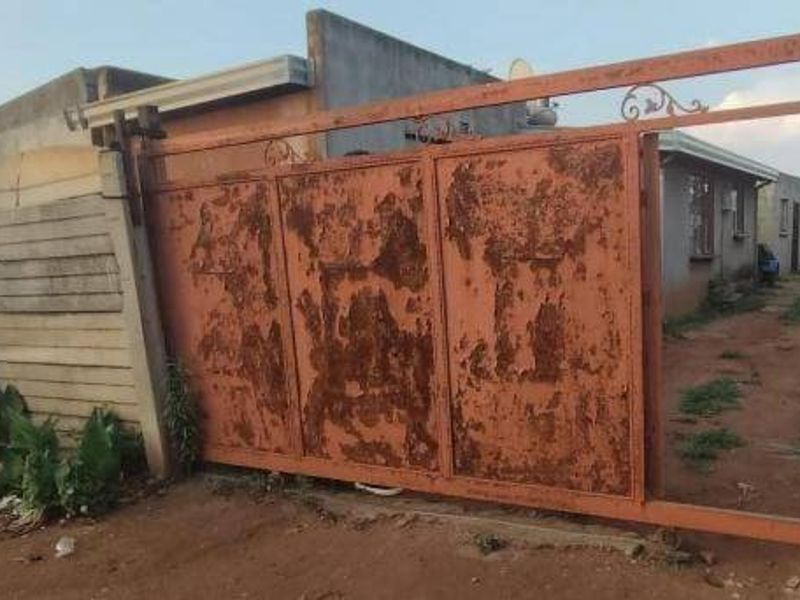 Front view of the property featuring a large sliding metal gate and precast concrete boundary wall, showing significant signs of wear and weathering.