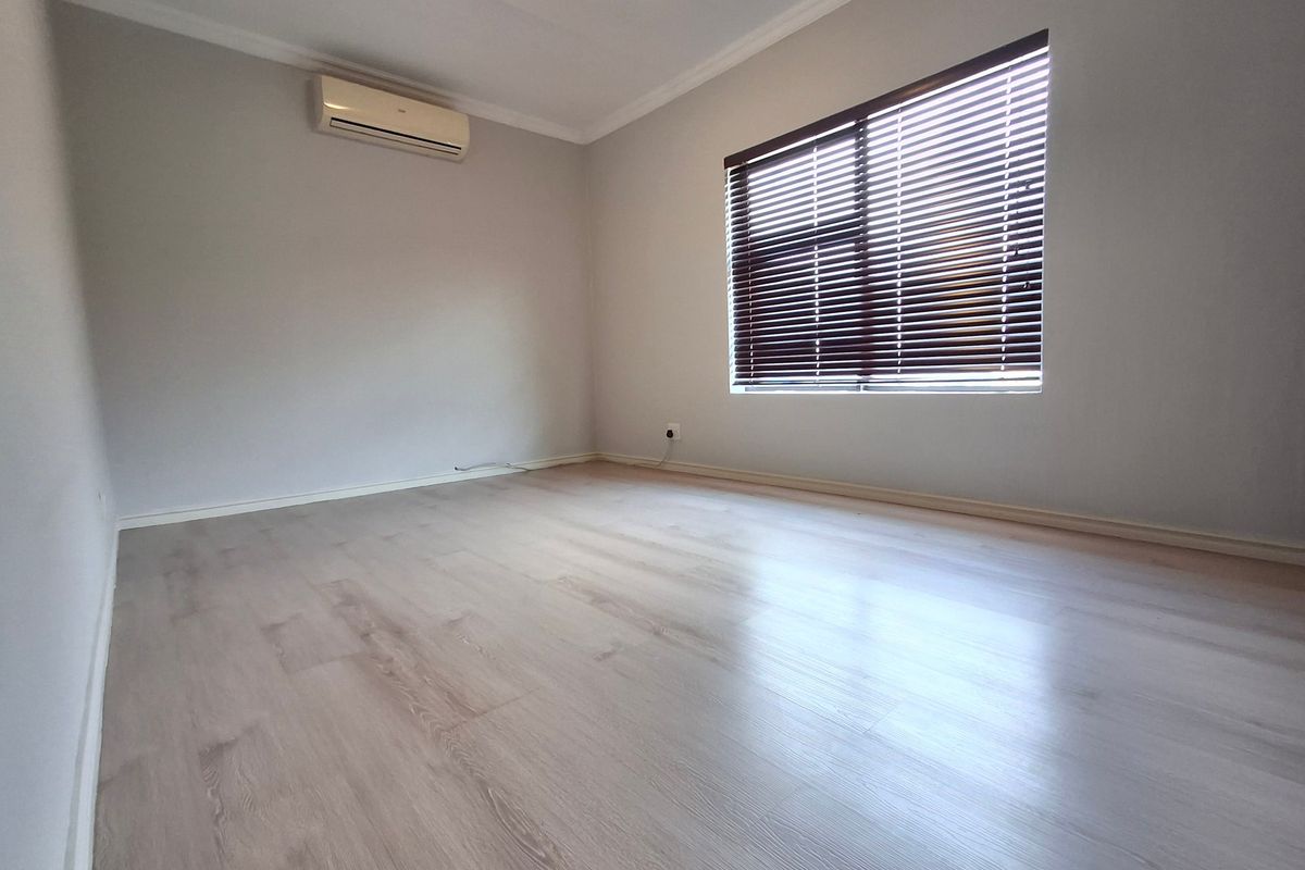 A bright, empty bedroom featuring light-toned laminate flooring, a wall-mounted air conditioning unit, and window blinds.