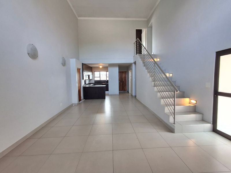 Spacious open-plan living area featuring high ceilings, tiled flooring, and a modern staircase with integrated lighting, leading to a compact kitchen.