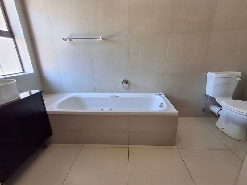 A clean, neutral-toned bathroom featuring a built-in bathtub, tiled flooring, wall tiling, and a standard toilet suite.