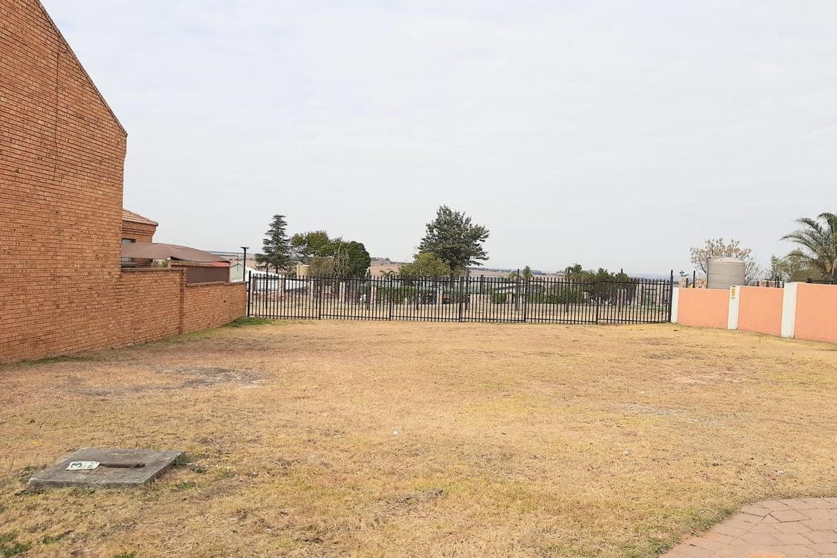 A vacant residential plot featuring a level grass area, enclosed by brick boundary walls and metal perimeter fencing with a visible water tank.