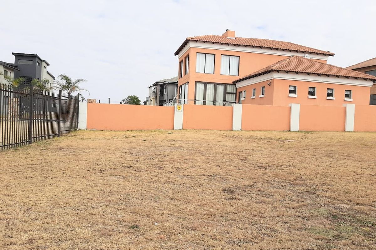 A view of a vacant residential plot featuring a perimeter wall and metal fencing, with neighboring residential properties visible in the background.