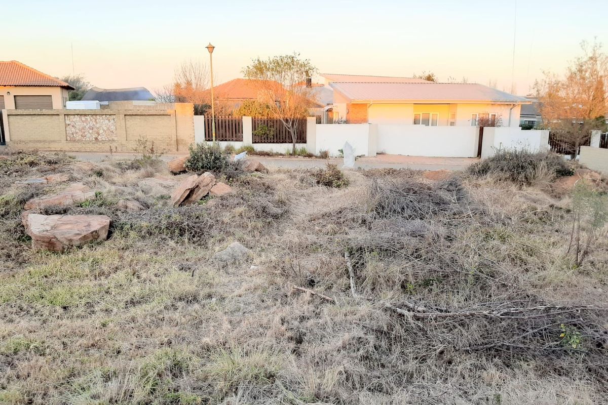 A vacant residential plot featuring natural rocky terrain and dry vegetation, situated in a developed neighborhood with established boundary walls.