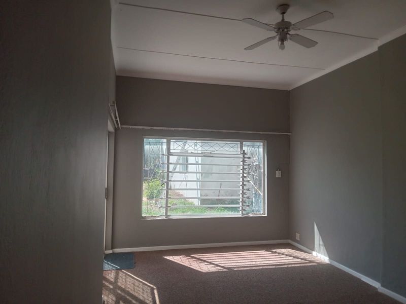 An empty living room featuring grey walls, carpeted flooring, a ceiling fan, and a louver window with security bars.