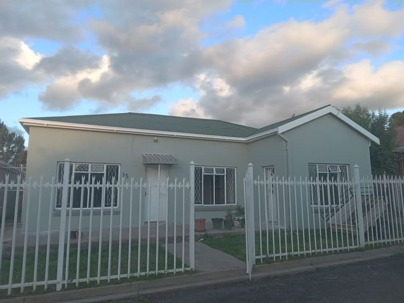 Front elevation of a single-story house featuring a metal roof, security fencing, and window bars, set behind a paved entrance area.