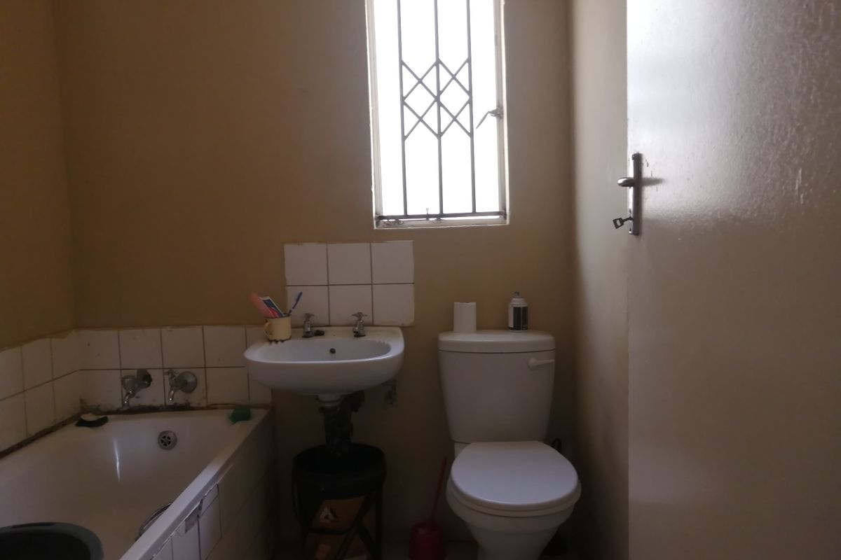 A functional bathroom featuring a standard bathtub, pedestal basin, and toilet, with security bars on the window and tiled splashback areas.