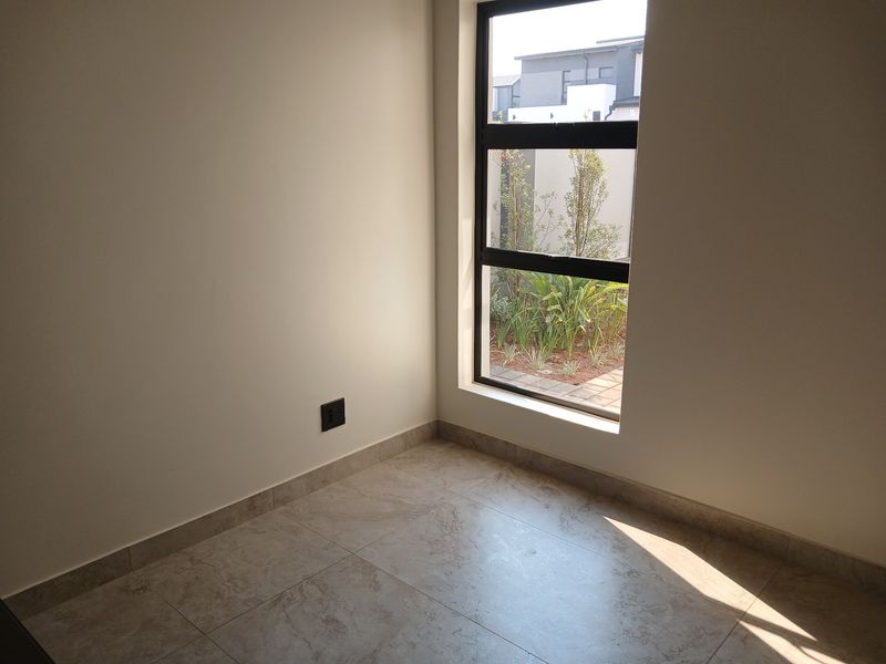 A bright, empty bedroom featuring neutral tiled flooring and a large window providing natural light and a view of the garden.