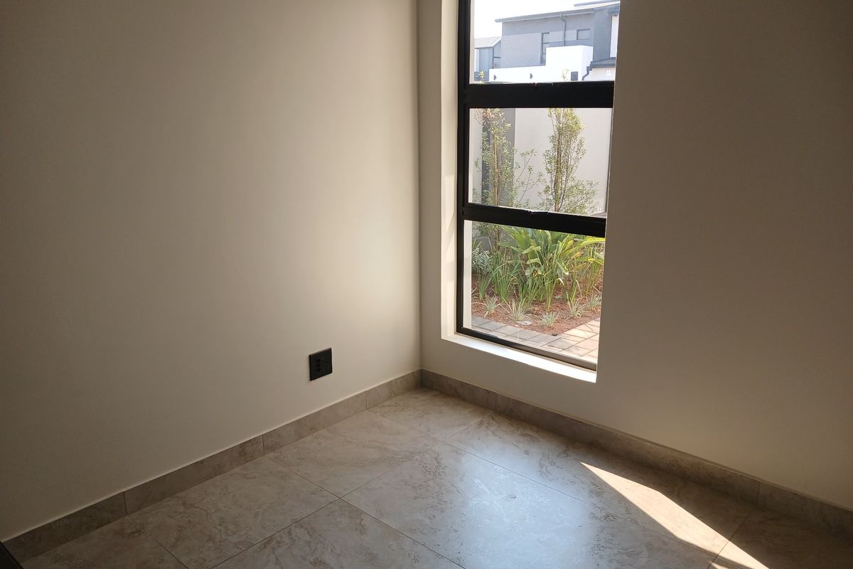 A bright, empty bedroom featuring neutral tiled flooring and a large window providing natural light and a view of the garden.