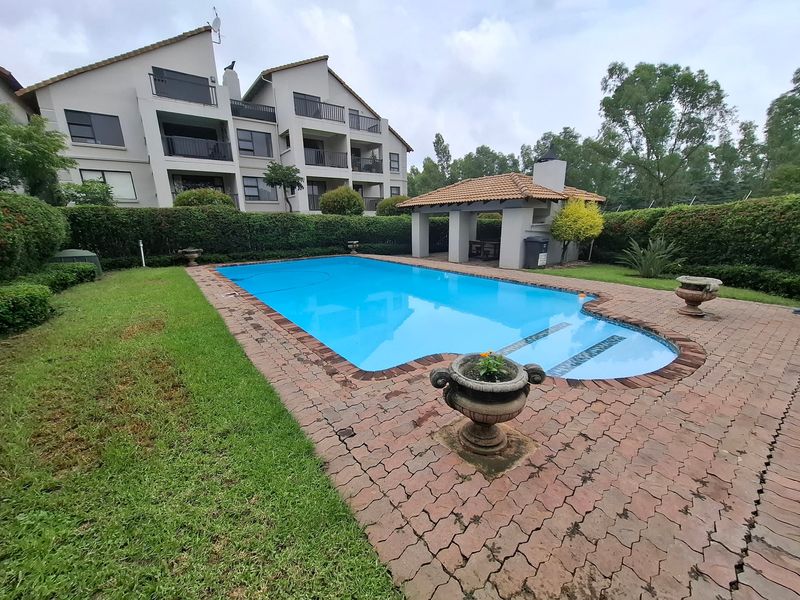 Communal swimming pool area featuring a covered gazebo, built-in braai, and brick paved surround set within a landscaped apartment complex garden.