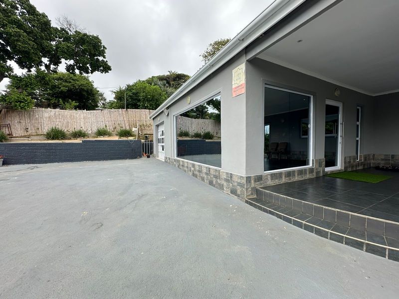 Front exterior view of the property featuring a paved driveway, a covered tiled patio, and large glass windows with a retaining wall in the background.