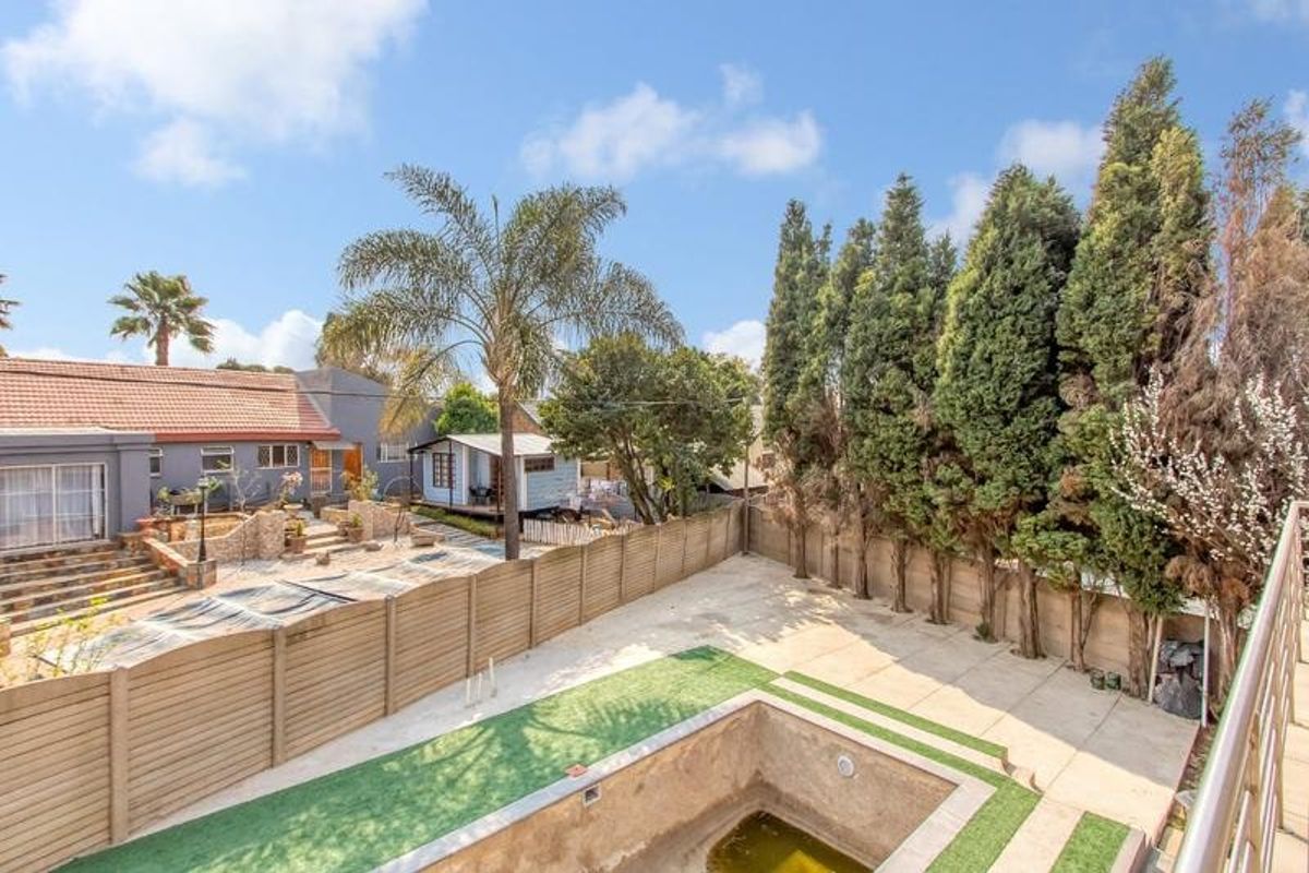 Rear exterior view featuring a paved patio area, artificial lawn, and an empty swimming pool with perimeter timber fencing and mature trees.