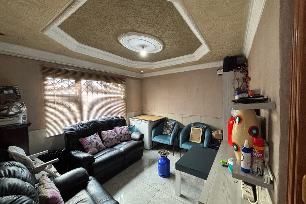 A living room featuring tiled flooring, a decorative tray ceiling with a central light fixture, and window blinds.