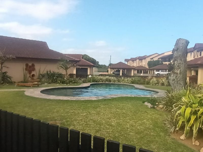 A communal outdoor pool area featuring a landscaped garden, lawn, and surrounding apartment complex buildings with tiled roofs.