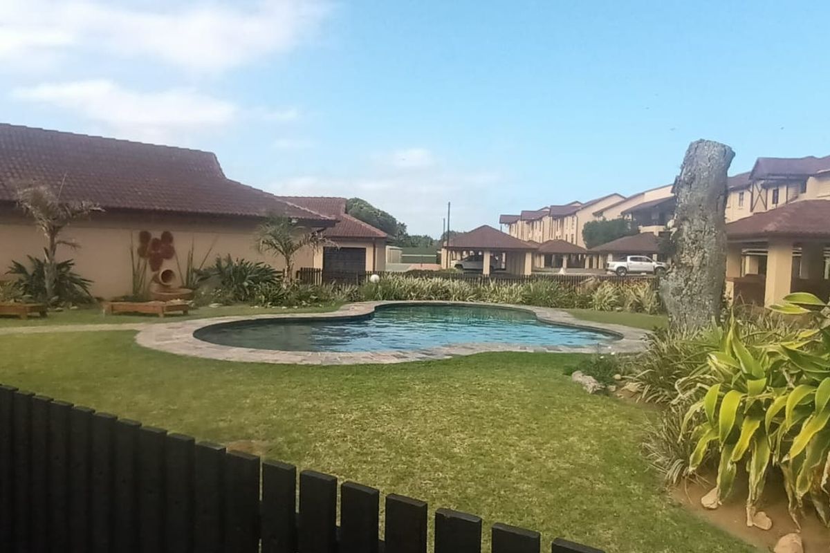 A communal outdoor pool area featuring a landscaped garden, lawn, and surrounding apartment complex buildings with tiled roofs.