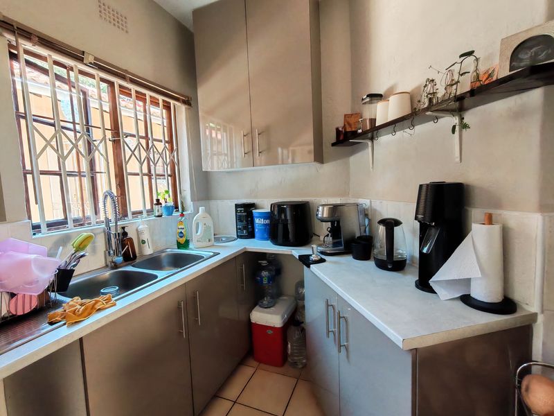 A compact kitchen featuring modern grey cabinetry, laminate countertops, a double stainless steel sink, and tiled finishes with security bars on the window.