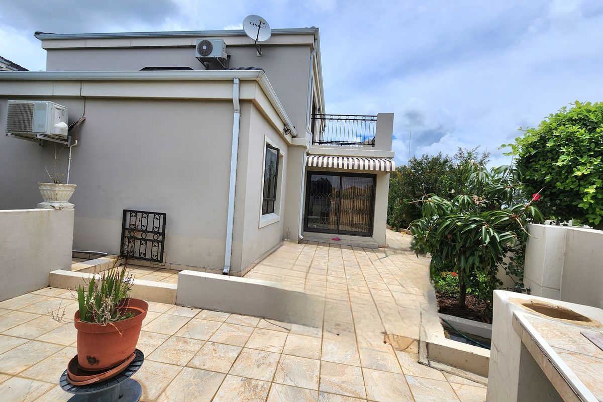 Rear exterior view featuring a tiled patio, built-in braai area, retractable awning, and an upper-level balcony with wrought iron railings.