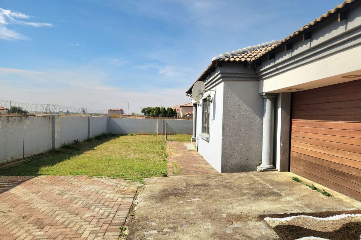 Rear exterior view featuring a paved driveway, brick patio area, perimeter wall with security fencing, and access to the garage.