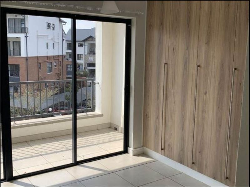 A bright bedroom featuring modern wood-finish built-in cupboards, tiled flooring, and a sliding glass door leading to a private balcony.