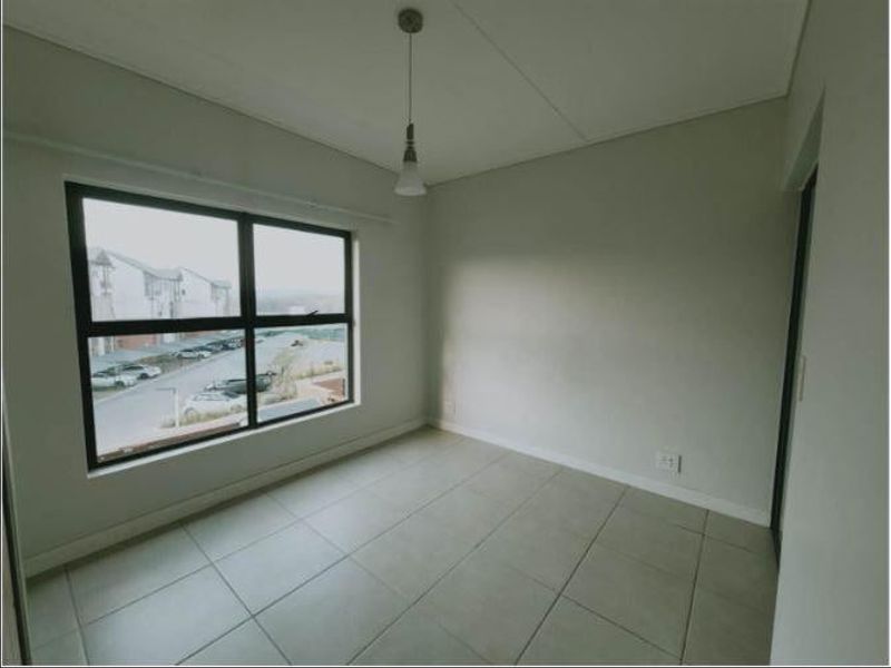 A bright, empty bedroom featuring neutral tiled flooring, a modern pendant light, and large windows providing natural light and a view of the complex.