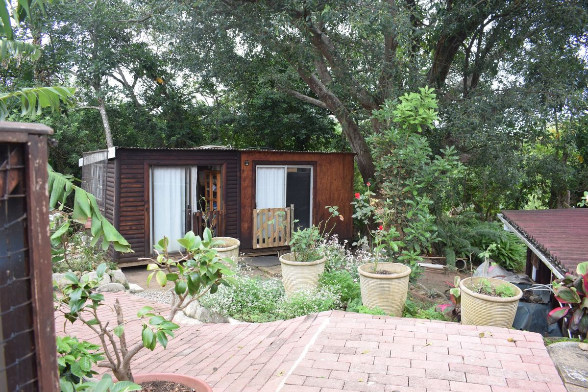 A rear view of a timber-clad structure set within a lush, mature garden, featuring a paved patio area and sliding glass door access.