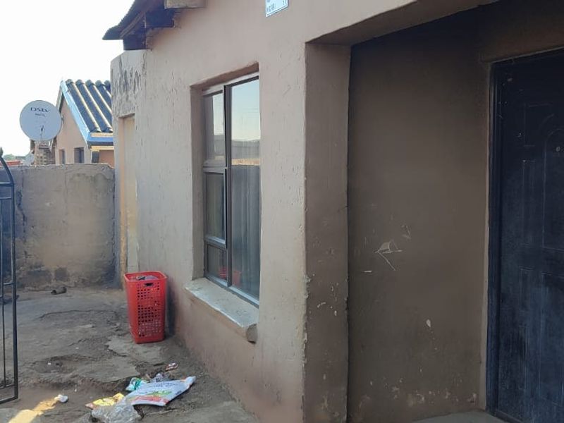 Front exterior view of the property featuring a metal security gate, solid entrance door, and aluminium window frame with visible wear to the wall finish.