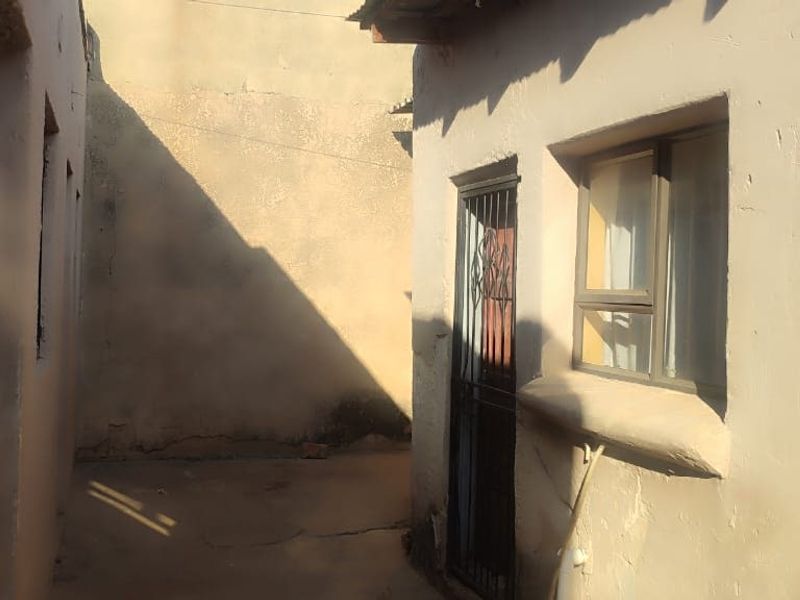 A narrow exterior side passage featuring concrete paving, a security gate, and an aluminium window frame, showing signs of weathered exterior plaster.